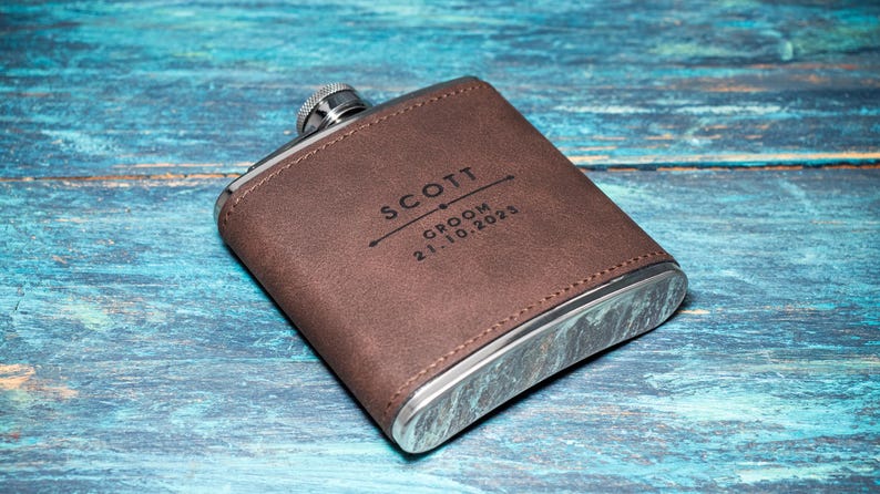 May include: A brown leather-covered flask with a silver cap and trim. The flask is personalized with the name "SCOTT", the word "GROOM", and the date "21.10.2025" in black lettering. The flask is set against a blue wooden background.