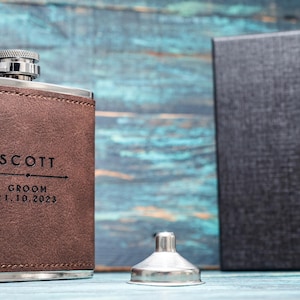 May include: A brown leather flask with silver accents, engraved with "SCOTT," "GROOM," and the date 21.10.2023. A silver funnel and a black box are also present. The flask is likely a gift.