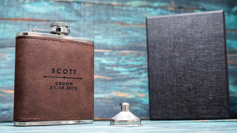 May include: A brown leather flask with silver accents, engraved with "SCOTT," "GROOM," and the date 21.10.2023. A silver funnel and a black box are also present. The flask is likely a gift.