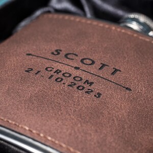 May include: A brown leather flask with the name "SCOTT" and the date "21.10.2023" engraved on the front. The flask is in a black box, suggesting a gift set. The flask has a silver rim.