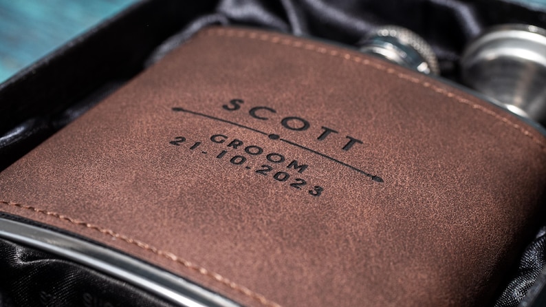 May include: A brown leather flask with the name "SCOTT" and the date "21.10.2023" engraved on the front. The flask is in a black box, suggesting a gift set. The flask has a silver rim.