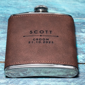 May include: A brown leather-wrapped flask with a silver cap and base. The flask is engraved with the name "SCOTT", the word "GROOM", and the date "21.10.2023". The flask is a rectangular shape.