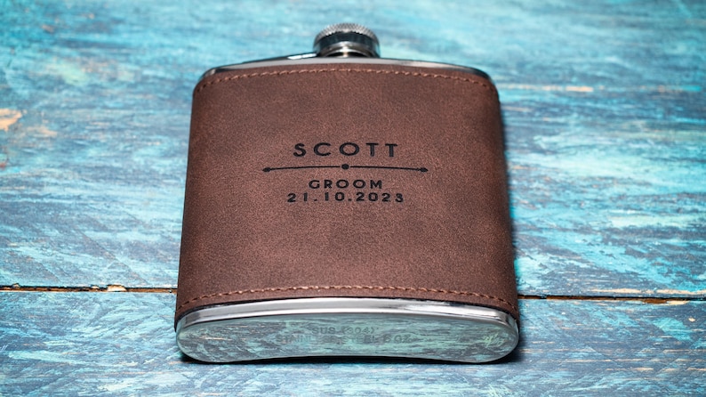 May include: A brown leather-wrapped flask with a silver cap and base. The flask is engraved with the name "SCOTT", the word "GROOM", and the date "21.10.2023". The flask is a rectangular shape.