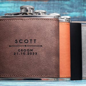 May include: A collection of personalized flasks in brown, orange, black, and gray leather. The front flask is engraved with "SCOTT", "GROOM", and the date "21.10.2023". Each flask has a silver cap.