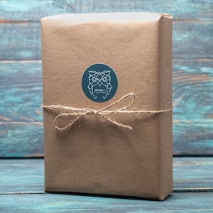May include: A rectangular package wrapped in brown paper, tied with twine, and sealed with a round sticker. The sticker features a geometric owl design and the text "WONBLY DESIGNS". The package is set against a blue and teal wooden background.