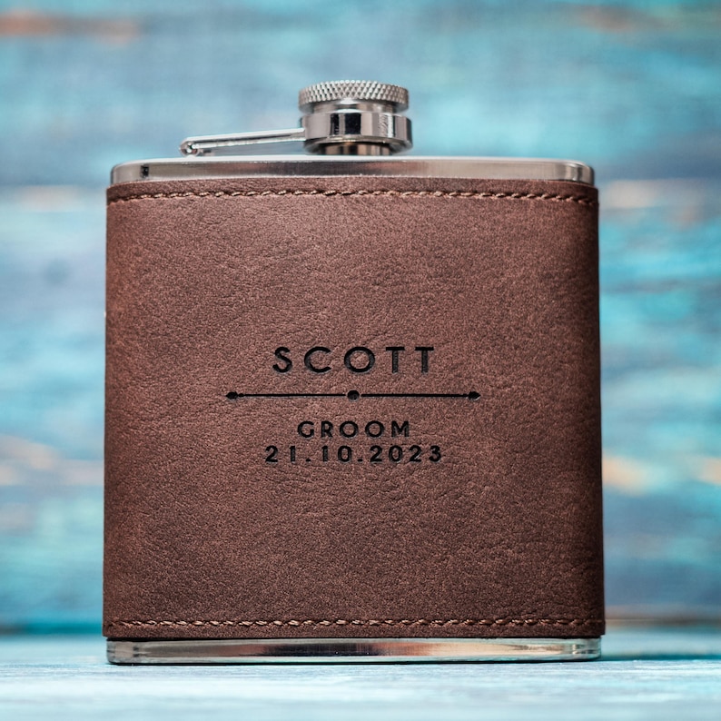 May include: A brown leather-wrapped flask with a silver cap and spout. The flask is engraved with the name "SCOTT", the word "GROOM", and the date "21.10.2023". The flask has a rectangular shape and is made of stainless steel.
