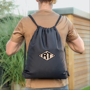 Könnte beinhalten: Schwarzer Kordelzugrucksack mit einem rautenförmigen Logo mit den Buchstaben "RT" und den Zahlen "19" und "89". Der Rucksack hat schwarze Kordelzüge und einen Reißverschluss. Die Person trägt ein braunes T-Shirt.