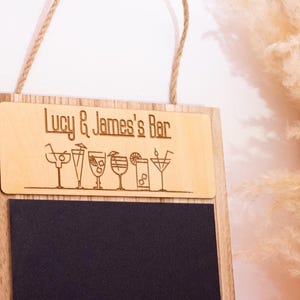May include: A wooden sign with a chalkboard insert. The top part of the sign is made of light wood and has the text "Lucy & James's Bar" engraved on it. There are also small drawings of cocktails on the sign. The bottom part of the sign is a black chalkboard.