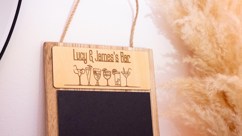 May include: A wooden sign with a chalkboard insert. The top part of the sign is made of light wood and has the text "Lucy & James's Bar" engraved on it. There are also small drawings of cocktails on the sign. The bottom part of the sign is a black chalkboard.