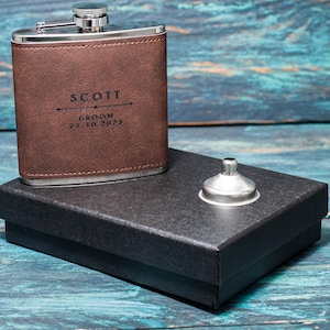 May include: A brown leather flask with a silver cap, engraved with the name "SCOTT", the word "GROOM", and the date "21.10.2023". It sits on a black box with a silver funnel. The background is a blue wooden surface.