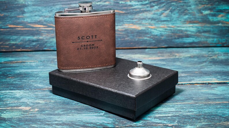 May include: A brown leather flask with a silver cap, engraved with the name "SCOTT", the word "GROOM", and the date "21.10.2023". It sits on a black box with a silver funnel. The background is a blue wooden surface.