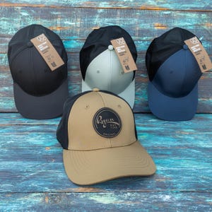 May include: Four baseball caps with mesh backs and solid front panels in black, light grey, navy blue, and tan. The tan cap features a circular black patch with the text Ryan EST. 1986 ORIGINAL. Each cap includes a brown cardboard hang tag.
