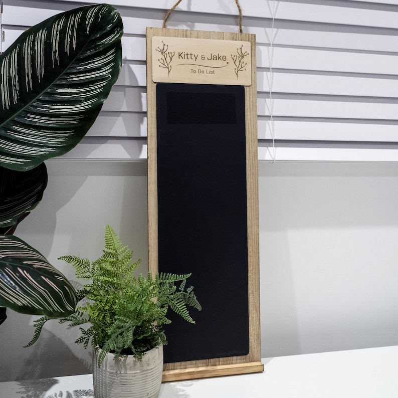 May include: A wooden to-do list board with a chalkboard surface. The board has a wooden frame and a hanging rope. The top of the board has a wooden sign with the text "Kitty & Jake To Do List" engraved on it.