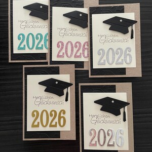 Peut inclure: Plusieurs cartes de remise de diplômes avec des coiffes noires. Chaque carte affiche l'année "2026" dans diverses couleurs, dont le turquoise, le rose, l'argent et l'or, avec le texte allemand "Herzlichen Glückwunsch". Les cartes ont un fond noir et beige texturé.