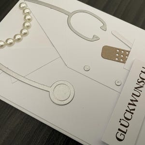 Könnte beinhalten: Eine weiße Grußkarte mit einem silberfarbenen Stethoskop-Stanzteil, einem kleinen braunen Pflaster und einer Reihe weißer Perlen. Auf der Vorderseite befindet sich ein weißer Papierstreifen mit dem schwarzen Aufdruck GLÜCKWUNSCH.
