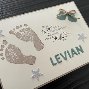 Peut inclure: Carte crème avec deux empreintes de pas de bébé en encre taupe, des étoiles argentées et le nom "LEVIAN" en lettres vertes. La carte comporte également du texte en allemand et un petit nœud.
