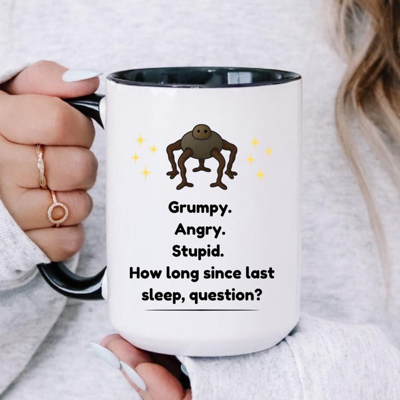 May include: A white ceramic mug with a black interior and handle. The mug features the words 'Grumpy. Angry. Stupid. How long since last sleep, question?' in black text, along with a cartoon image of a brown creature with yellow star accents.