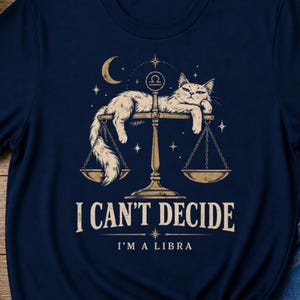 May include: A navy blue t-shirt featuring a graphic of a fluffy cat lounging on a set of golden scales. Above the cat is a crescent moon and the Libra zodiac symbol. Below the scales, the text reads I CAN'T DECIDE I'M A LIBRA in a serif font.
