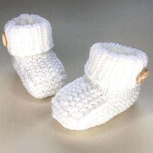 Puede incluir: Un par de botines de bebé blancos tejidos. Cada botín tiene un puño doblado y un pequeño botón de madera. Los botines están hechos de un tejido grueso y están diseñados para mantener los pies del bebé calientes.