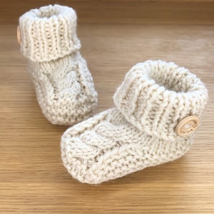 May include: A pair of cream-colored knitted baby booties with a wooden button on each. The button on the right bootie has the text "With love" and a heart symbol.