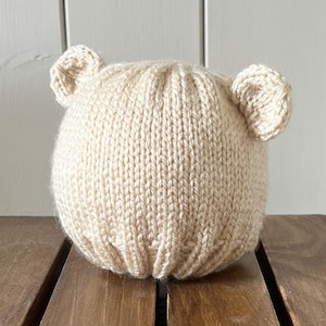 May include: A beige knitted hat with two small, rounded ears. The hat is made of a thick, textured yarn and sits on a wooden surface. The hat has a round shape and is designed to fit a baby or toddler.