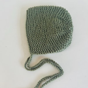 May include: A knitted olive green baby bonnet with attached ties. The bonnet is made of a textured yarn and has a rounded crown. The ties are made of the same yarn and are designed to be tied under the chin.
