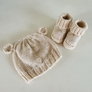 Op de afbeelding: Een beige gebreide baby set bestaande uit een muts met twee kleine oren en een paar bijpassende babyslofjes. De set is gemaakt van zacht, getextureerd garen, ideaal voor een pasgeborene. De muts en slofjes worden op een licht oppervlak getoond.