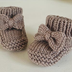 Może przedstawiać: Para dzianinowych bucików dla niemowląt w miękkim brązowym kolorze. Każdy bucik ma dekoracyjną kokardę. Buciki wykonane są z teksturowanej przędzy, idealne dla noworodka. Mają około 8 cm długości.