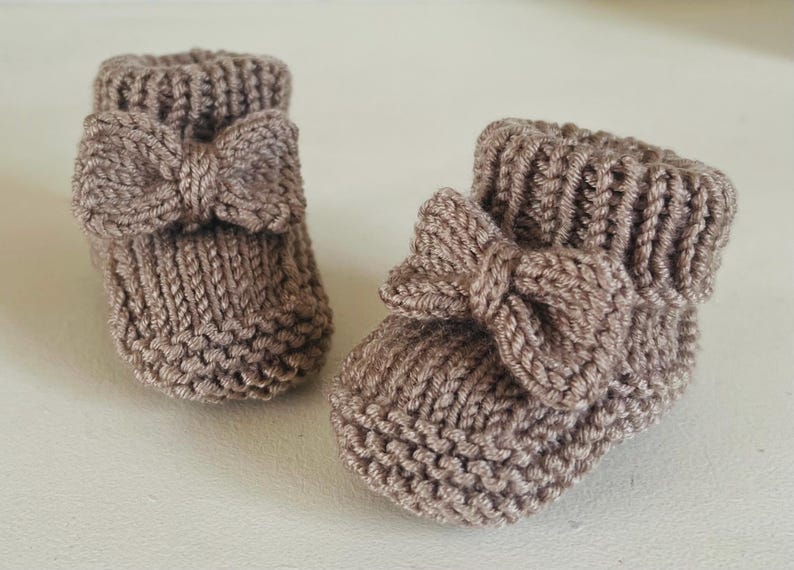 May include: Two brown knitted baby booties with bow accents. The booties are made of a soft, textured yarn and feature a folded cuff. The bows are centered on the front of each bootie, adding a decorative touch.