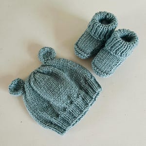 Op de afbeelding: Een gebreide baby set in een zachte blauwgroene kleur. De set bevat een muts met twee kleine oor-achtige details en een paar bijpassende babyslofjes. De items zijn gemaakt van een dikke gebreide stof.