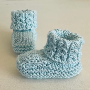 Pode incluir: Um par de botinhas de bebê de malha azul claro. As botinhas têm uma bainha dobrada com um padrão texturizado. O fio parece macio e as botinhas são projetadas para um recém-nascido. As botinhas são feitas à mão.