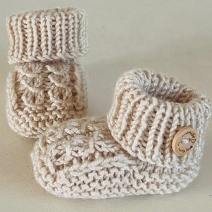 Könnte beinhalten: Ein Paar cremefarbene Strickschühchen mit geripptem Bündchen und einem strukturierten Zopfmuster am Körper. Ein Schühchen ist mit einem kleinen, runden Holzknopf verziert, der mit dem Text "handmade with love" und einem Herzsymbol graviert ist.