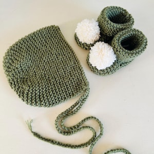 Può includere: Un set per neonati lavorato a maglia in verde oliva, composto da un cappellino con lacci e un paio di scarpine. Le scarpine hanno pompon bianchi. Il set è realizzato in filato morbido, perfetto per un neonato.