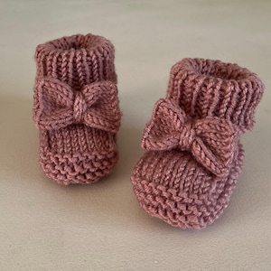 Puede incluir: Un par de patucos de bebé tejidos en color rosa empolvado. Cada patuco presenta un lazo decorativo en la parte delantera. Los patucos están hechos de un hilo suave y texturizado, ideal para recién nacidos.