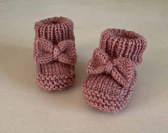 May include: A pair of knitted baby booties in a dusty rose color. Each bootie features a decorative bow on the front. The booties are made of a soft, textured yarn, perfect for a newborn.
