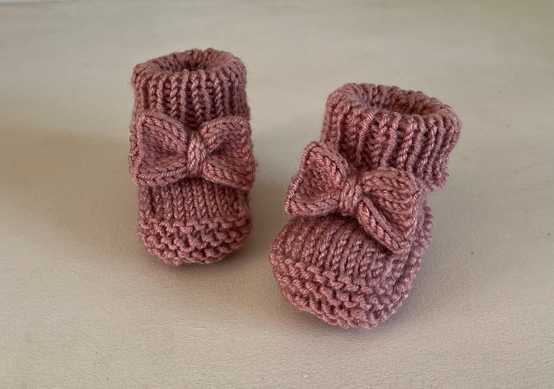 May include: A pair of knitted baby booties in a dusty rose color. Each bootie features a decorative bow on the front. The booties are made of a soft, textured yarn, perfect for a newborn.