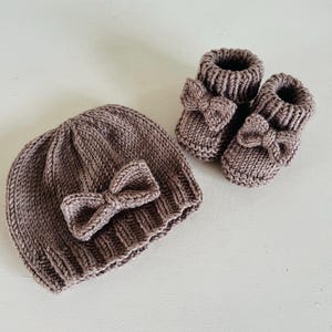 Può includere: Un set per neonati lavorato a maglia in un tenue colore malva, composto da un berretto e un paio di stivaletti. Ogni articolo è adornato da un fiocco decorativo. Il set è realizzato in filato morbido, perfetto per un neonato.