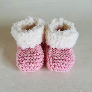 Könnte beinhalten: Ein Paar Baby-Booties, gestrickt in hellrosa Garn. Sie haben einen flauschigen, cremefarbenen Besatz am oberen Rand. Diese Booties sind entworfen, um die Füße eines Babys warm zu halten.