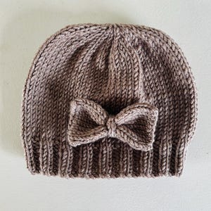 May include: A knitted beanie in a muted brown color, featuring a decorative bow on the front. The hat is made of a textured yarn, with a ribbed brim and a rounded crown. The bow is also knitted and adds a touch of detail to the design.