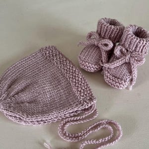 May include: A knitted baby set in a soft mauve color, including a bonnet with ties and a pair of booties with bow details. The set is made of a textured yarn, perfect for a newborn.