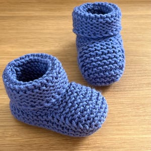 Op de afbeelding: Twee handgebreide blauwe babyslofjes. De slofjes zijn gemaakt van zacht, getextureerd garen en hebben een opgerolde manchet aan de bovenkant. Ze zijn ontworpen om de voetjes van een baby warm te houden. Gepresenteerd op een houten oppervlak.