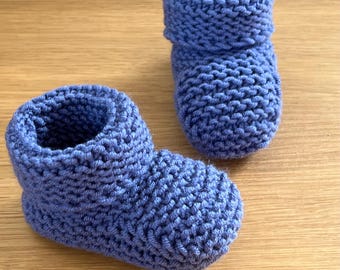 May include: Two handmade, blue knitted baby booties. The booties are made of a soft, textured yarn and have a rolled cuff at the top. They are designed to keep a baby's feet warm and cozy. The booties are displayed on a wooden surface.