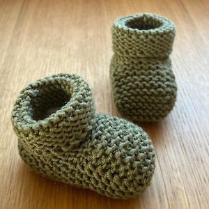 Può includere: Due scarpine da neonato lavorate a maglia verde oliva. Le scarpine sono realizzate con un filato spesso e hanno un polsino a coste. Una scarpina è sdraiata su un lato, mentre l'altra è in posizione verticale. Le scarpine sono su una superficie di legno marrone chiaro.
