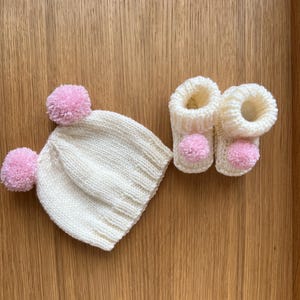 Pode incluir: Um conjunto de bebé de malha com um chapéu branco com dois pompons cor-de-rosa e um par de botinhas a condizer com pompons cor-de-rosa. O conjunto é feito de fio macio e texturizado, perfeito para um recém-nascido. Os artigos são apresentados numa superfície de madeira.