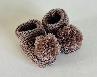 May include: A pair of brown crochet baby booties with fluffy pom-poms. The booties are made of a textured yarn and have a soft, cozy appearance. The pom-poms add a playful touch to the design.