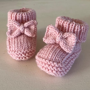 May include: A pair of pale pink knitted baby booties. Each bootie has a bow on the front and a ribbed cuff. The booties are made of soft yarn and are designed for a newborn.