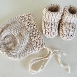 May include: A cream-colored knitted baby bonnet and matching booties. The bonnet has a decorative border and ties. The booties have a ribbed cuff and button detail. The set is made of a soft, textured yarn.