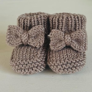 May include: A pair of knitted baby booties in a soft taupe color. Each bootie features a decorative bow on the front. The booties are made of a textured yarn and have a ribbed cuff.