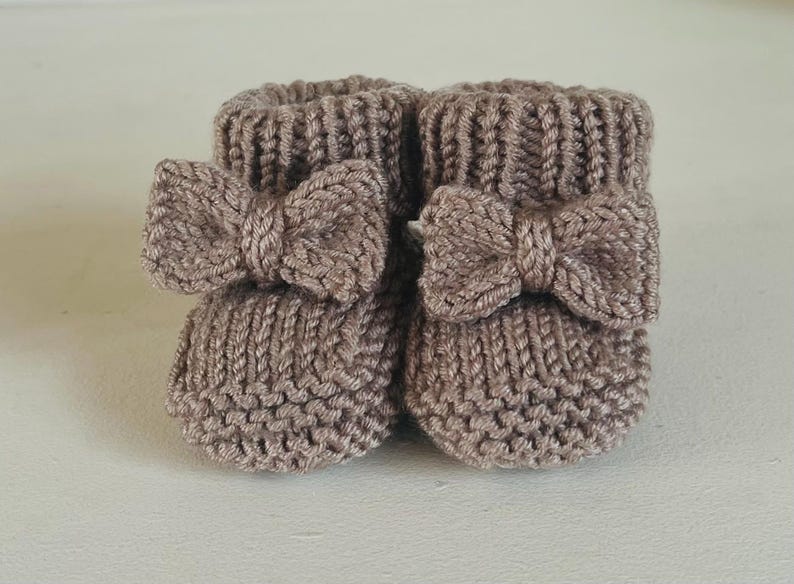 May include: A pair of knitted baby booties in a soft taupe color. Each bootie features a decorative bow on the front. The booties are made of a textured yarn and have a ribbed cuff.