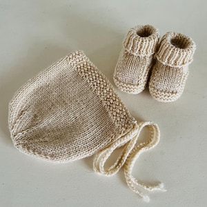 Może przedstawiać: Dzianinowy zestaw dla dziecka w neutralnym beżowym kolorze. Zestaw zawiera czapeczkę ze sznurkami do wiązania i parę bucików. Elementy wykonane są z miękkiej, teksturowanej przędzy.